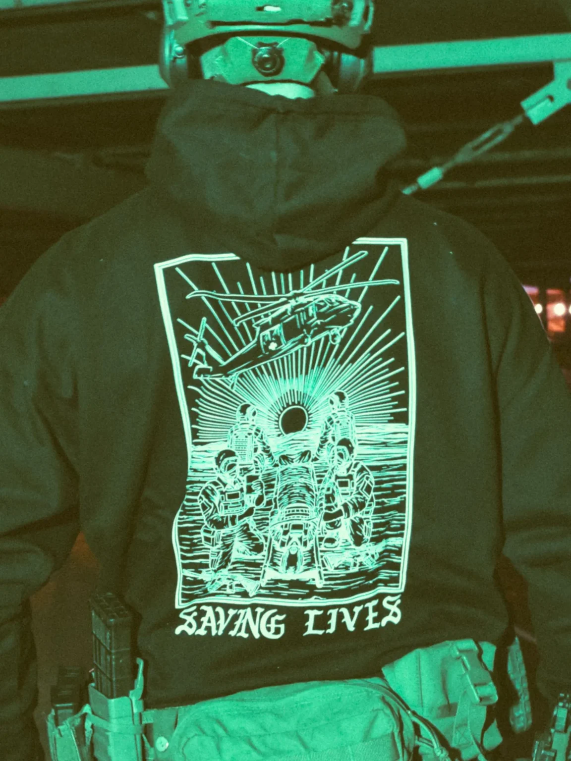 Saving-Lives-hoodie-black-edit Military-style hoodie featuring the Saving Lives design: a group of soldiers carrying a stretcher while a helicopter prepares to land, symbolizing courage under pressure.