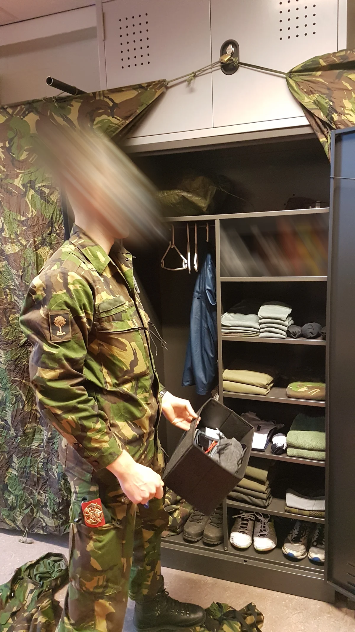 Dutch airmobile recruit beside his locker during training, kit folded and squared away, camouflage net draped over the top