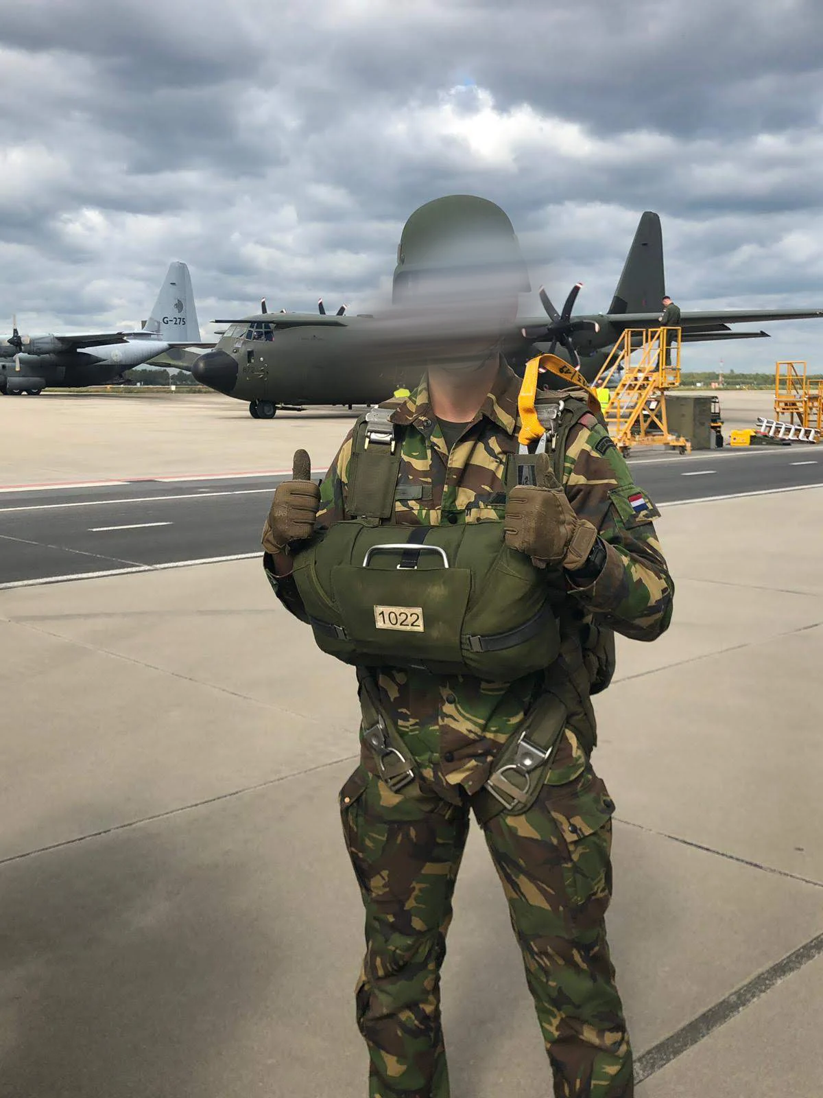 Young Dutch paratrooper rigged with a parachute and reserve giving a thumbs up, C-130 Hercules on the apron behind him