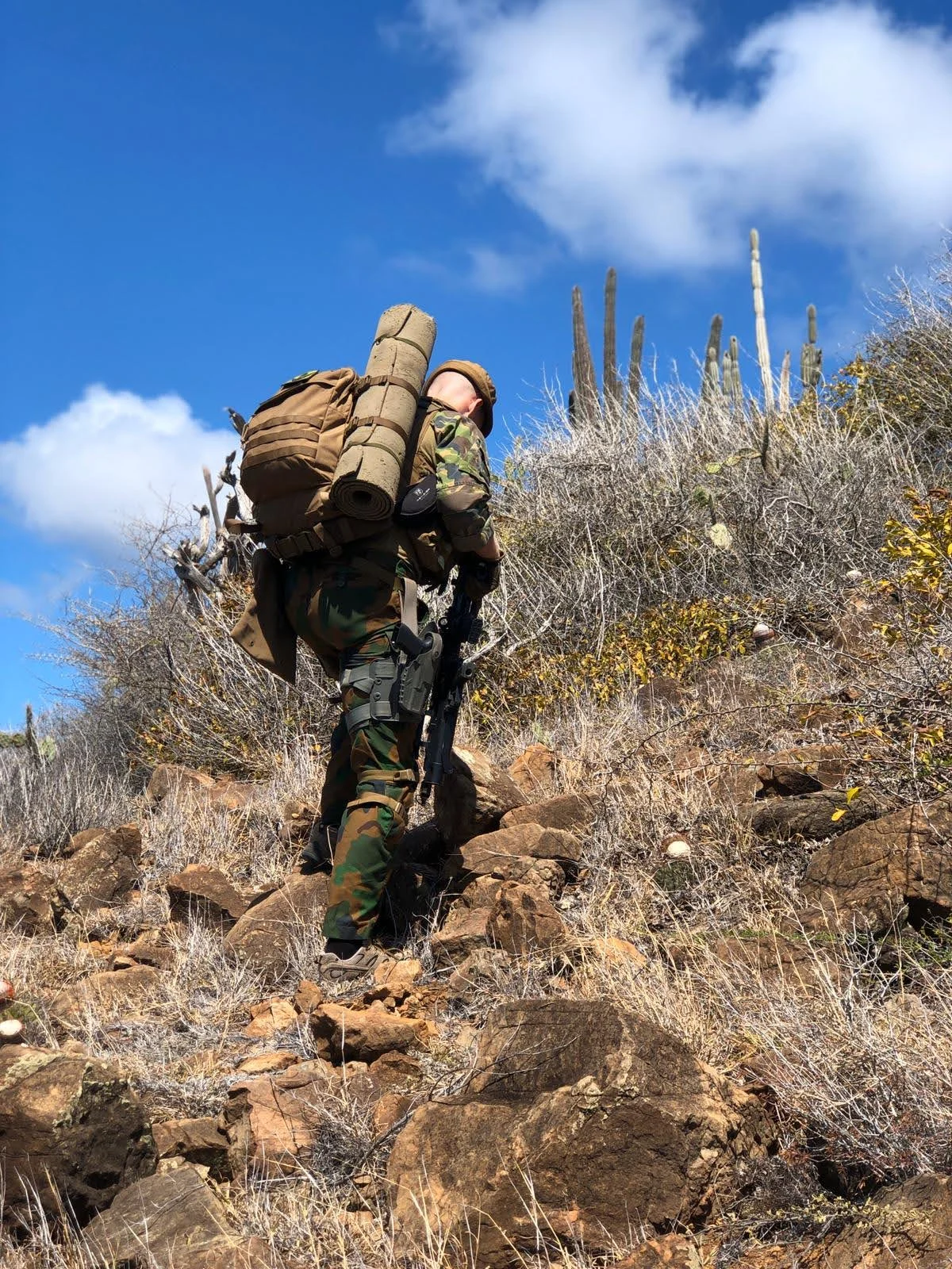Dutch soldier climbing a rocky cactus covered hillside in Curacao carrying a full pack and roll mat