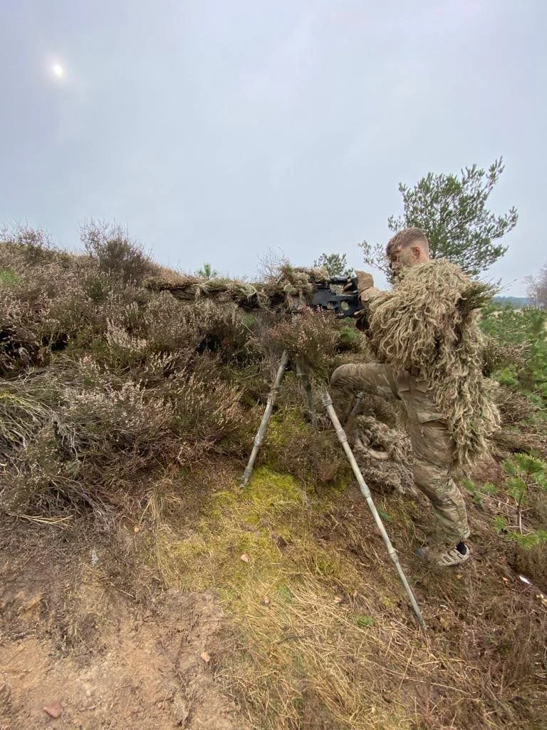Sniper in a ghillie suit behind a rifle on a tripod in heathland