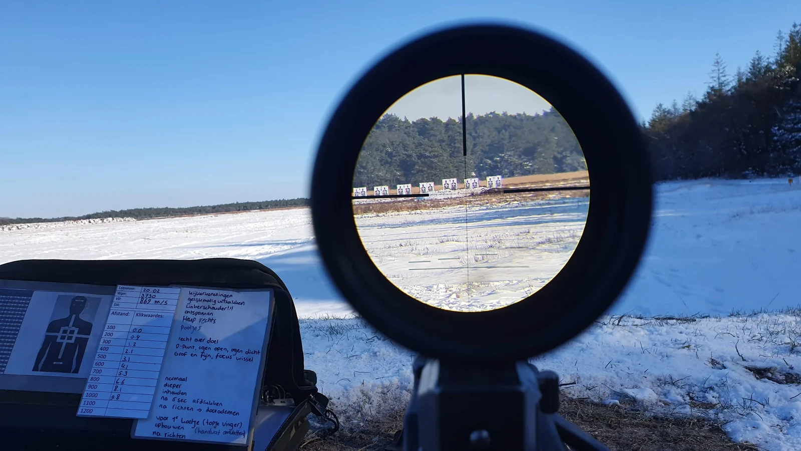 View through a rifle scope at targets on a snow covered range, handwritten range data card in the foreground
