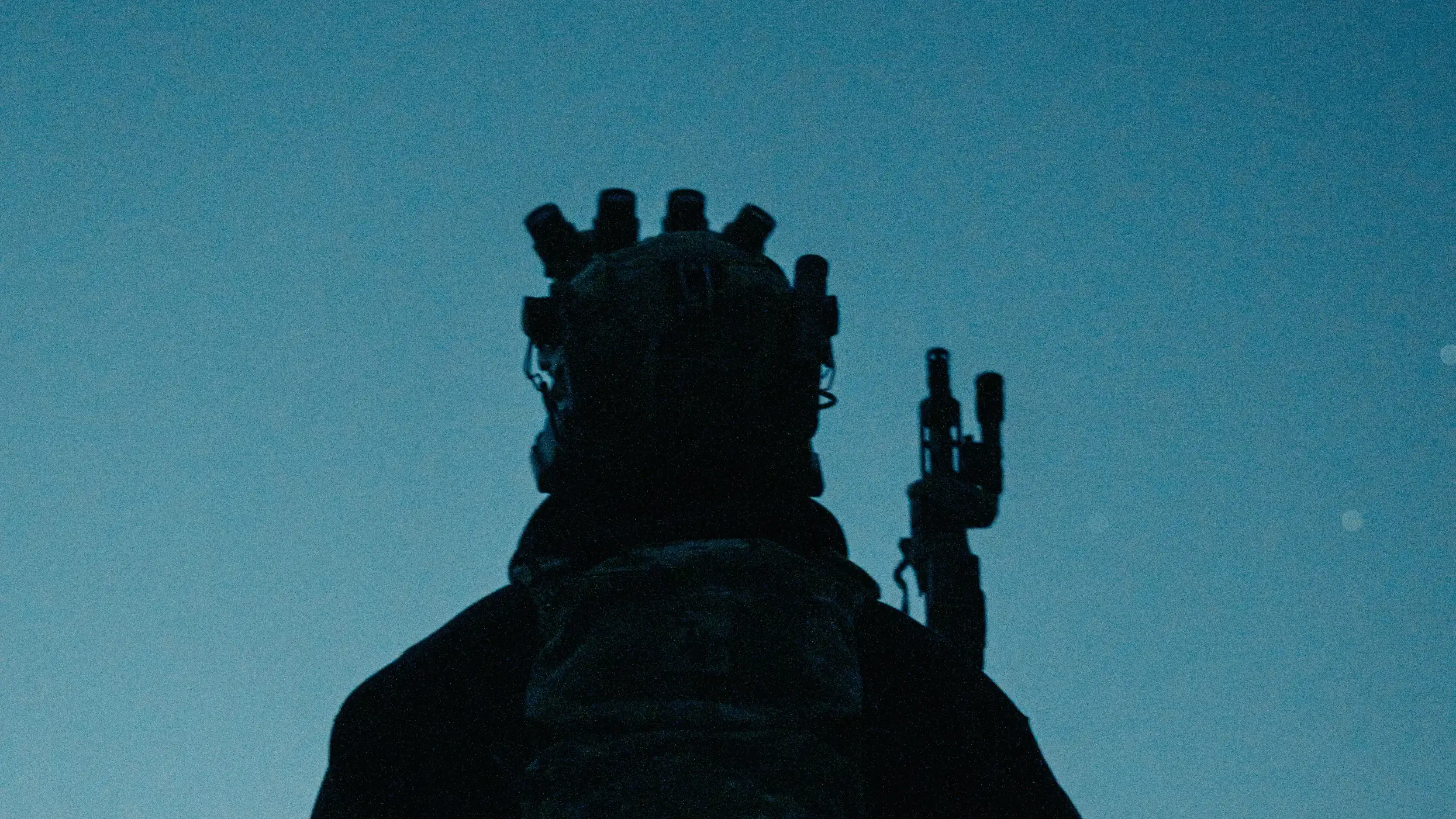 Operator seen from behind at blue hour, four tube night vision on the helmet and rifle raised