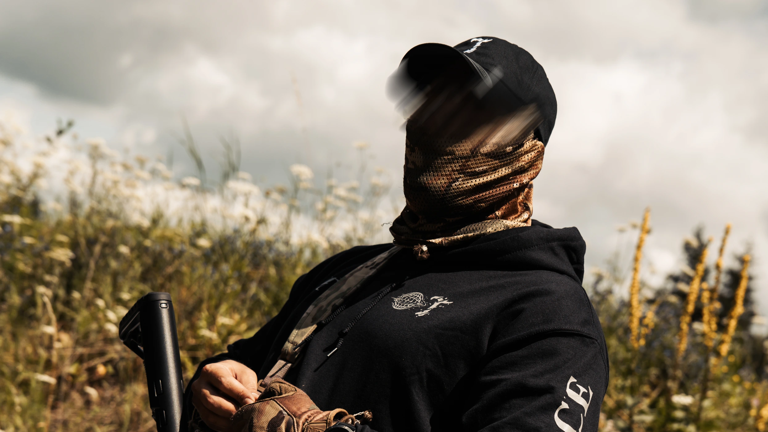 The Trace black hoodie worn by an operator with shemagh and cap in an open field