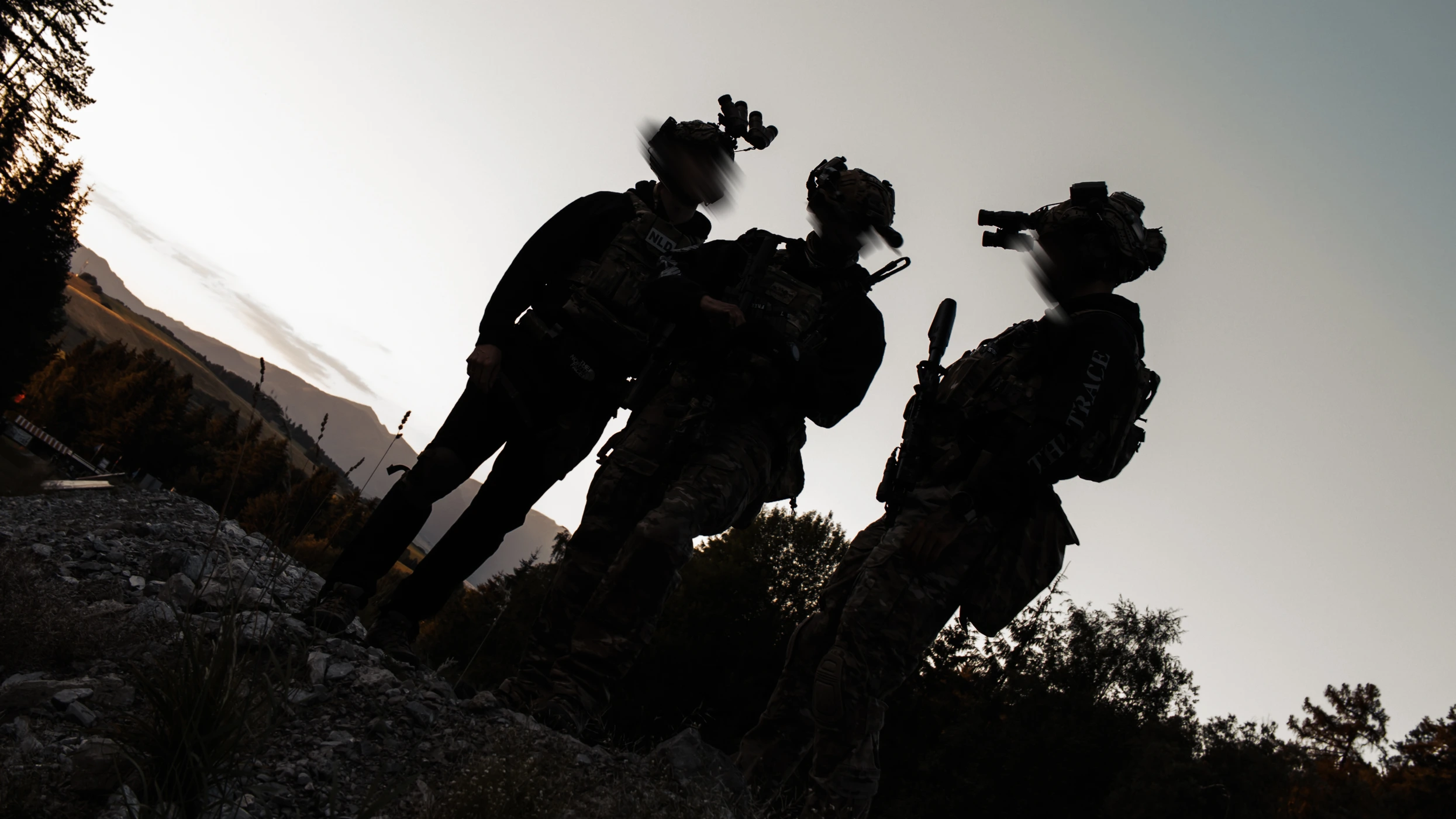 Three operators silhouetted against a bright sky on a ridge, night vision on their helmets